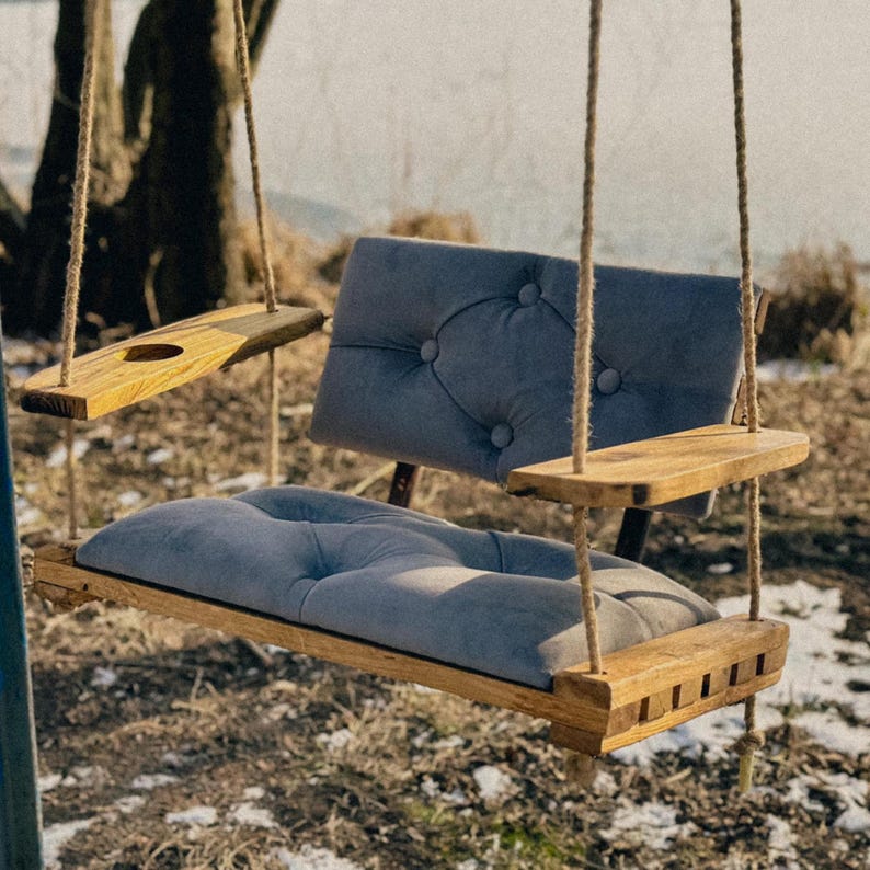May include: A wooden swing with a gray cushioned seat and a small wooden shelf on the side. The swing is hanging from two ropes.