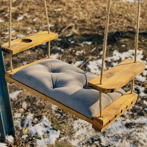 May include: A wooden swing with a gray cushioned seat and two wooden armrests. The swing is hanging from ropes attached to a metal frame.