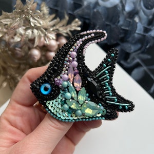 May include: A black beaded brooch in the shape of a fish with a turquoise and green iridescent body, a black eye with a blue pupil, and pink and purple accents.