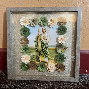 May include: A framed picture of a religious figure with a green robe and a staff, surrounded by paper flowers in shades of green, white, and brown. The background is a silver glitter paper.