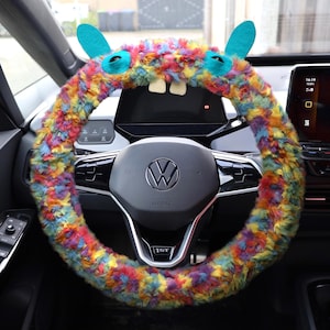 May include: A colorful, fuzzy steering wheel cover in a car. The cover is decorated with blue ears, eyes, and two white teeth, giving it a monster-like appearance. The cover is made of a mix of rainbow-colored fur.