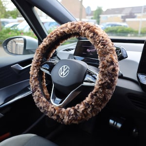 Pode incluir: Uma capa de volante de pele sintética marrom e preta. A capa está instalada em um volante preto com um logotipo da Volkswagen prateado.