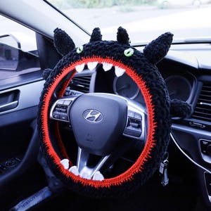 Pu&ograve; includere: Un coprivolante in uncinetto nero a forma di creatura con occhi verdi, orecchie a punta e una fodera interna rossa con denti triangolari bianchi. La copertura &egrave; montata su un volante nero con il logo Hyundai argentato al centro.