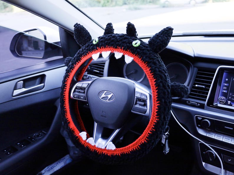 Pu&ograve; includere: Un coprivolante in uncinetto nero a forma di creatura con occhi verdi, orecchie a punta e una fodera interna rossa con denti triangolari bianchi. La copertura &egrave; montata su un volante nero con il logo Hyundai argentato al centro.