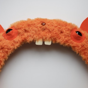 Puede incluir: Una diadema de felpa naranja con textura peluda, dos orejas de fieltro naranja erguidas, dos ojos circulares oscuros sobre parches de fieltro naranja y dos dientes blancos prominentes que sobresalen del centro de la estructura curva.