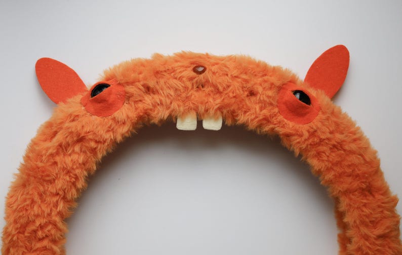 Puede incluir: Una diadema de felpa naranja con textura peluda, dos orejas de fieltro naranja erguidas, dos ojos circulares oscuros sobre parches de fieltro naranja y dos dientes blancos prominentes que sobresalen del centro de la estructura curva.