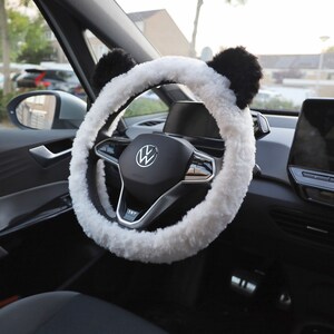 May include: White faux fur steering wheel cover with black panda ears. The cover is installed on a black steering wheel with a silver Volkswagen logo.