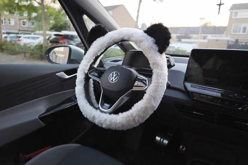 May include: White faux fur steering wheel cover with black panda ears. The cover is installed on a black steering wheel with a silver Volkswagen logo.