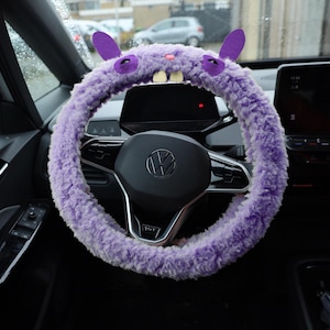 Puede incluir: Una funda de volante morada y peluda con un dise&ntilde;o de criatura que incluye dos orejas erguidas, ojos bordados en morado oscuro, una peque&ntilde;a nariz rosa y dos dientes blancos prominentes que sobresalen del centro del tejido de felpa.