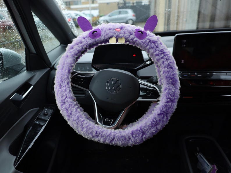 Puede incluir: Una funda de volante morada y peluda con un dise&ntilde;o de criatura que incluye dos orejas erguidas, ojos bordados en morado oscuro, una peque&ntilde;a nariz rosa y dos dientes blancos prominentes que sobresalen del centro del tejido de felpa.