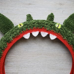Pu&ograve; includere: Un copricapo a forma di mostro realizzato all'uncinetto con un esterno in filato verde scuro testurizzato, una fodera interna arancione brillante, quattro denti bianchi appuntiti e due occhi gialli con pupille verticali nere, incluse orecchie.