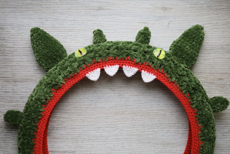 Pu&ograve; includere: Un copricapo a forma di mostro realizzato all'uncinetto con un esterno in filato verde scuro testurizzato, una fodera interna arancione brillante, quattro denti bianchi appuntiti e due occhi gialli con pupille verticali nere, incluse orecchie.