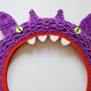 Pu&ograve; includere: Una cornice circolare a forma di mostro realizzata all'uncinetto in viola con una lavorazione in filato spesso e testurizzato. Il design presenta due occhi verde brillante con pupille nere, piccole corna appuntite e denti bianchi.