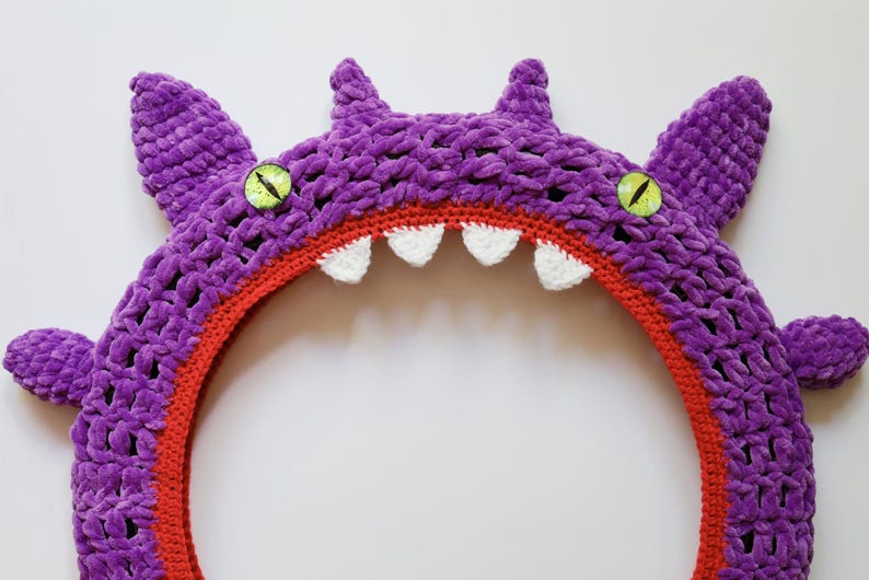 Pu&ograve; includere: Una cornice circolare a forma di mostro realizzata all'uncinetto in viola con una lavorazione in filato spesso e testurizzato. Il design presenta due occhi verde brillante con pupille nere, piccole corna appuntite e denti bianchi.