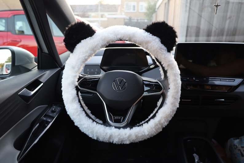 May include: White fluffy steering wheel cover with black panda ears. The cover is installed on a black steering wheel with a silver Volkswagen logo.