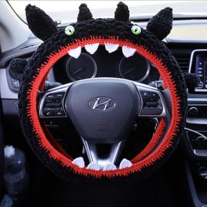 May include: A black crocheted steering wheel cover shaped like a creature with green eyes, pointed ears, and a red inner lining featuring white triangular teeth. The cover is fitted over a black steering wheel with a silver Hyundai logo in the center.