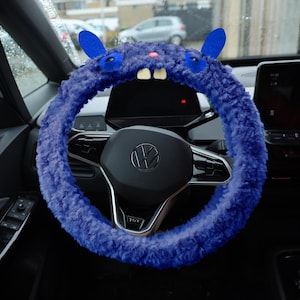 Puede incluir: Una funda para volante de color morado intenso y textura peluda con forma de monstruo, que presenta dos orejas azules, dos ojos oscuros, una peque&ntilde;a nariz rosa y dos dientes blancos prominentes sobre un volante negro con el logo de Volkswagen.