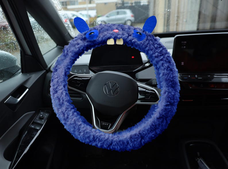 Puede incluir: Una funda para volante de color morado intenso y textura peluda con forma de monstruo, que presenta dos orejas azules, dos ojos oscuros, una peque&ntilde;a nariz rosa y dos dientes blancos prominentes sobre un volante negro con el logo de Volkswagen.