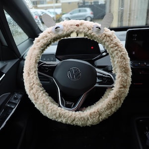 Puede incluir: Una funda para volante de felpa beige y peluda que presenta dos orejas erguidas, dos ojos circulares y dos dientes rectangulares blancos en la parte superior. La funda est&aacute; instalada en un volante negro con el logo de Volkswagen en el centro.