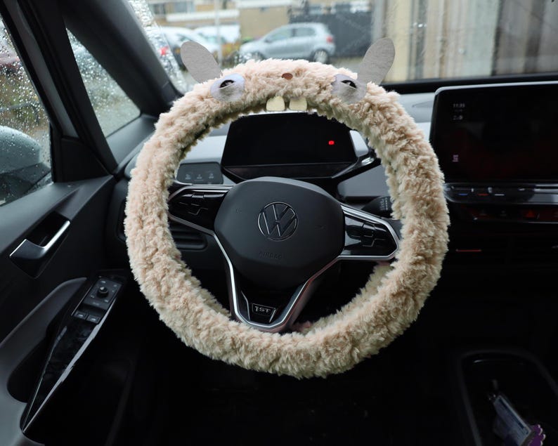 Puede incluir: Una funda para volante de felpa beige y peluda que presenta dos orejas erguidas, dos ojos circulares y dos dientes rectangulares blancos en la parte superior. La funda est&aacute; instalada en un volante negro con el logo de Volkswagen en el centro.