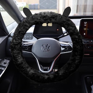Puede incluir: Una funda de volante de piel sint&eacute;tica negra con dos orejas erguidas en la parte superior, instalada en un volante Volkswagen con un logotipo central y detalles plateados, situada en el interior de un veh&iacute;culo con pantalla digital.
