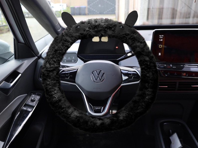 Puede incluir: Una funda de volante de piel sint&eacute;tica negra con dos orejas erguidas en la parte superior, instalada en un volante Volkswagen con un logotipo central y detalles plateados, situada en el interior de un veh&iacute;culo con pantalla digital.