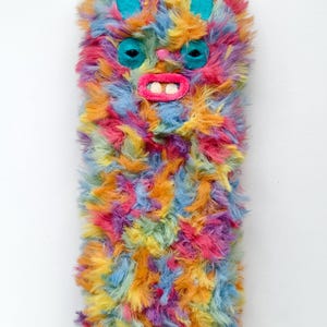 Puede incluir: Una funda rectangular para tel&eacute;fono cubierta de pelo sint&eacute;tico largo y multicolor en tonos rosa, naranja, amarillo y azul. La funda presenta la cara de una criatura con orejas de fieltro color cerceta, ojos oscuros, una peque&ntilde;a nariz rosa y una boca rosa brillante con dos dientes blancos.