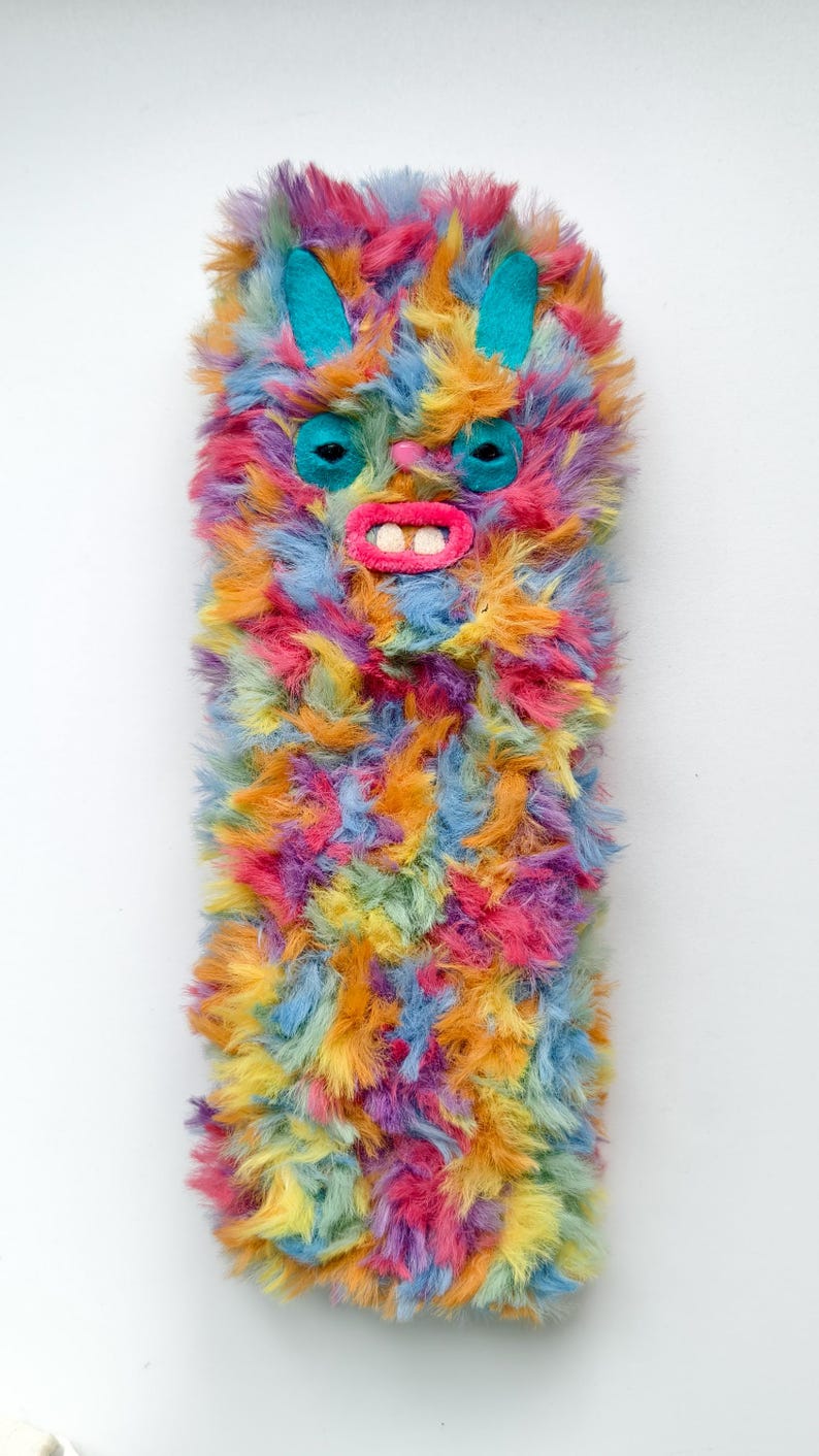 Puede incluir: Una funda rectangular para tel&eacute;fono cubierta de pelo sint&eacute;tico largo y multicolor en tonos rosa, naranja, amarillo y azul. La funda presenta la cara de una criatura con orejas de fieltro color cerceta, ojos oscuros, una peque&ntilde;a nariz rosa y una boca rosa brillante con dos dientes blancos.