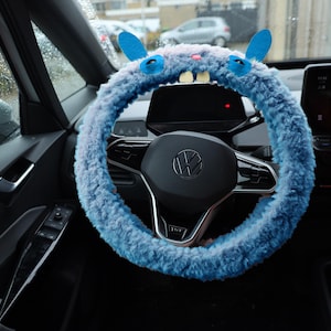Puede incluir: Una funda de volante de felpa azul claro con forma de criatura, con dos orejas erguidas, ojos de fieltro azul, una peque&ntilde;a nariz rosa y dos dientes blancos prominentes. La funda est&aacute; instalada en un volante negro con el logo de Volkswagen.