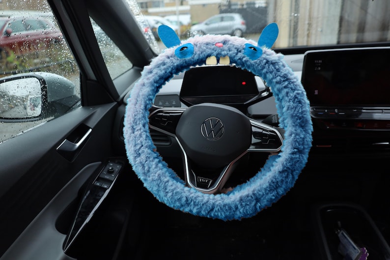 Puede incluir: Una funda de volante de felpa azul claro con forma de criatura, con dos orejas erguidas, ojos de fieltro azul, una peque&ntilde;a nariz rosa y dos dientes blancos prominentes. La funda est&aacute; instalada en un volante negro con el logo de Volkswagen.