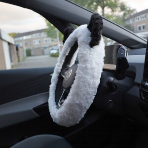 May include: White fuzzy steering wheel cover with black panda ears. The cover is installed on a black steering wheel in a car.