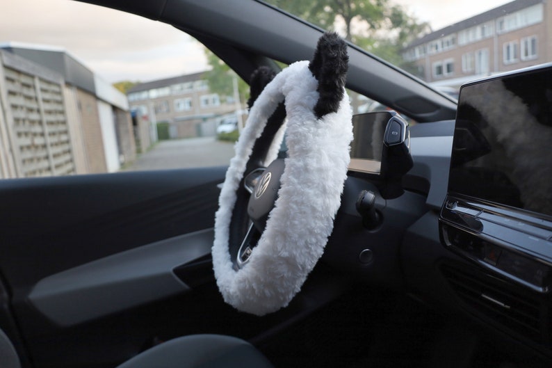 May include: White fuzzy steering wheel cover with black panda ears. The cover is installed on a black steering wheel in a car.