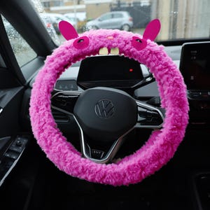Puede incluir: Una funda para volante de color rosa brillante y textura peluda, que presenta dos orejas erguidas, dos ojos oscuros y dos dientes blancos prominentes en la parte superior. El material afelpado envuelve el volante circular de un veh&iacute;culo, a&ntilde;adiendo una capa decorativa suave al componente de control interior.