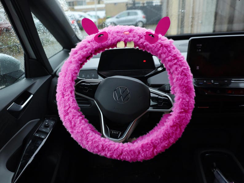 Puede incluir: Una funda para volante de color rosa brillante y textura peluda, que presenta dos orejas erguidas, dos ojos oscuros y dos dientes blancos prominentes en la parte superior. El material afelpado envuelve el volante circular de un veh&iacute;culo, a&ntilde;adiendo una capa decorativa suave al componente de control interior.
