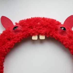 Puede incluir: Una diadema roja brillante y difusa que presenta dos orejas erguidas, dos ojos oscuros incrustados y dos dientes blancos prominentes que sobresalen del centro. La textura de la diadema es gruesa y afelpada, creando un aspecto suave y vibrante.