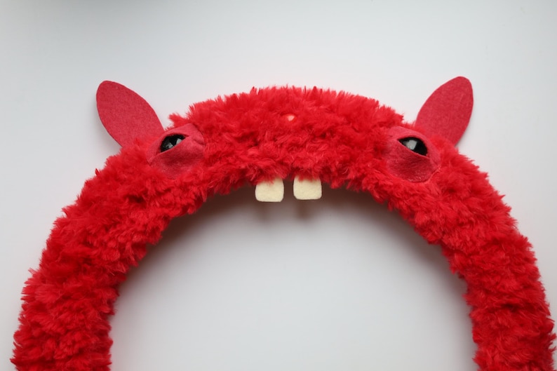 Puede incluir: Una diadema roja brillante y difusa que presenta dos orejas erguidas, dos ojos oscuros incrustados y dos dientes blancos prominentes que sobresalen del centro. La textura de la diadema es gruesa y afelpada, creando un aspecto suave y vibrante.