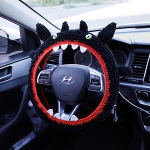 Pu&ograve; includere: Un coprivolante all'uncinetto nero progettato per assomigliare a una creatura con orecchie a punta, occhi verdi e una bocca spalancata con una fodera interna rosso brillante e denti triangolari bianchi affilati attorno al volante.