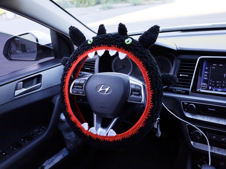 Pu&ograve; includere: Un coprivolante all'uncinetto nero progettato per assomigliare a una creatura con orecchie a punta, occhi verdi e una bocca spalancata con una fodera interna rosso brillante e denti triangolari bianchi affilati attorno al volante.
