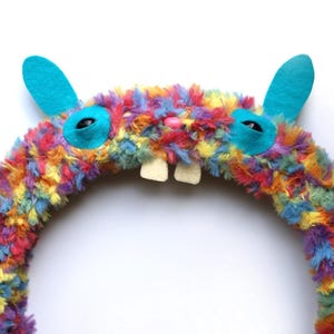 Puede incluir: Una corona circular cubierta de piel sint&eacute;tica esponjosa y multicolor en tonos naranja, amarillo, azul, morado y rojo. Presenta dos orejas de fieltro de color turquesa brillante, dos parches oculares turquesas con pupilas negras, una peque&ntilde;a nariz rosa y dos dientes blancos.