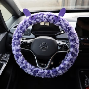 Puede incluir: Una funda de volante de peluche morado y blanco con dos orejas erguidas, dos ojos y dos dientes blancos. La funda est&aacute; instalada en un volante negro con un logotipo central de Volkswagen, situado en el interior de un veh&iacute;culo.