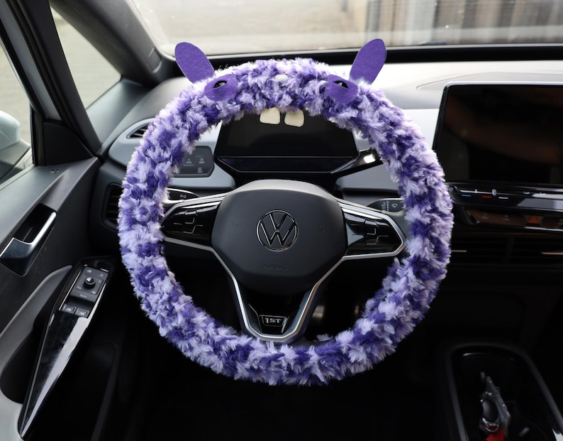Puede incluir: Una funda de volante de peluche morado y blanco con dos orejas erguidas, dos ojos y dos dientes blancos. La funda est&aacute; instalada en un volante negro con un logotipo central de Volkswagen, situado en el interior de un veh&iacute;culo.