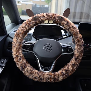Puede incluir: Una funda para volante de peluche en tonos marr&oacute;n y beige con dise&ntilde;o de animal, que incluye dos orejas erguidas, dos ojos oscuros y dos dientes blancos prominentes en la parte superior, instalada en un volante negro Volkswagen.