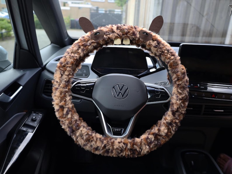 Puede incluir: Una funda para volante de peluche en tonos marr&oacute;n y beige con dise&ntilde;o de animal, que incluye dos orejas erguidas, dos ojos oscuros y dos dientes blancos prominentes en la parte superior, instalada en un volante negro Volkswagen.
