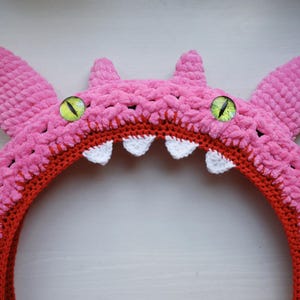 Pu&ograve; includere: Una fascia per capelli a forma di mostro lavorata all'uncinetto in rosa, con una superficie in filato testurizzato, due grandi occhi verdi con pupille verticali nere e una fila di piccoli denti triangolari bianchi lungo il bordo interno arancione.