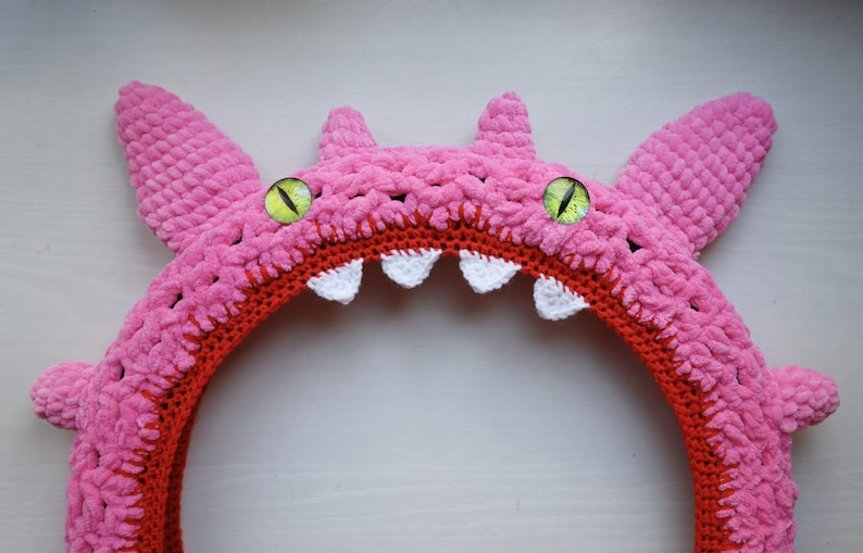 Pu&ograve; includere: Una fascia per capelli a forma di mostro lavorata all'uncinetto in rosa, con una superficie in filato testurizzato, due grandi occhi verdi con pupille verticali nere e una fila di piccoli denti triangolari bianchi lungo il bordo interno arancione.