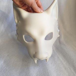 May include: A matte white, unpainted wolf mask featuring pointed ears, almond-shaped eye openings, and two prominent fangs extending downward from the upper jaw. The mask has a smooth, sculpted surface with defined snout and cheek details.