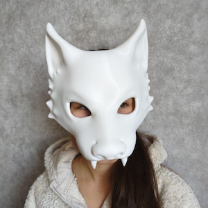 May include: A white wolf mask with pointed ears, a snout, and visible fangs. The mask has eye holes and is worn by a person. The mask is made of a smooth material and is a costume accessory.