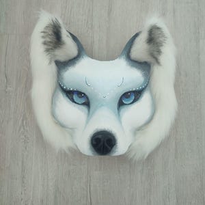 Pu&ograve; includere: Una maschera da lupo con dettagli dipinti in bianco e grigio, occhi blu brillante e un lungo bordo in pelliccia sintetica bianca attorno alle orecchie e ai lati. La maschera presenta un naso nero e delicati puntini bianchi sopra gli occhi.