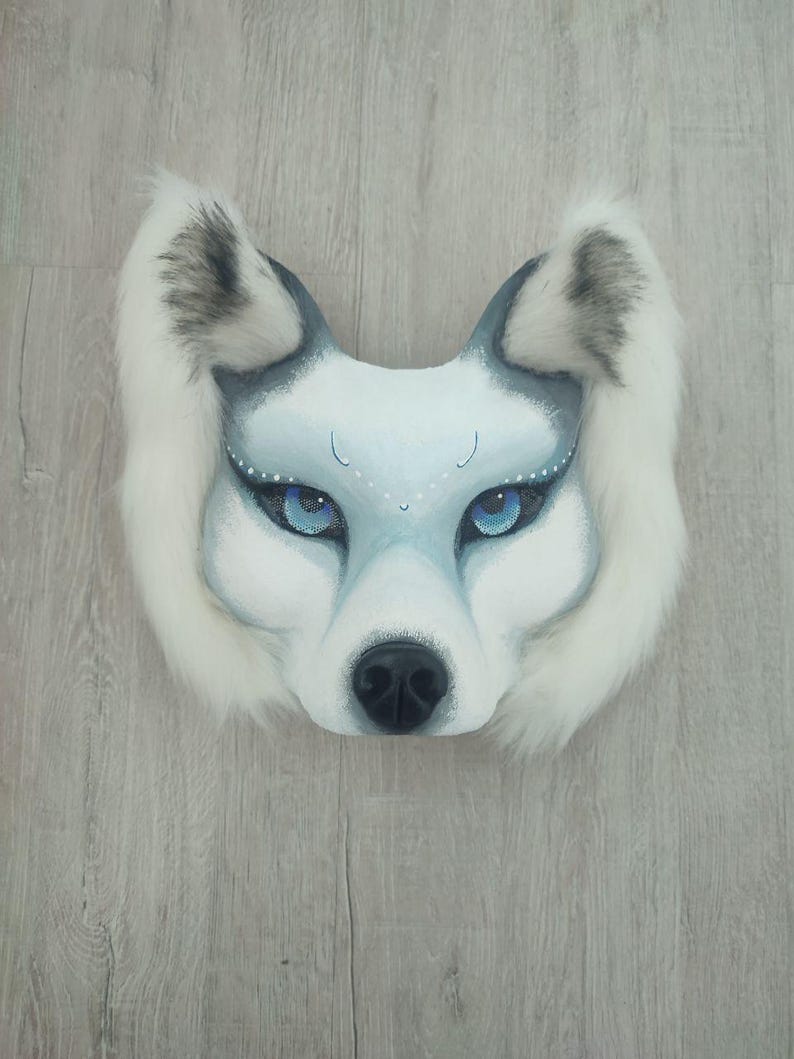Pu&ograve; includere: Una maschera da lupo con dettagli dipinti in bianco e grigio, occhi blu brillante e un lungo bordo in pelliccia sintetica bianca attorno alle orecchie e ai lati. La maschera presenta un naso nero e delicati puntini bianchi sopra gli occhi.