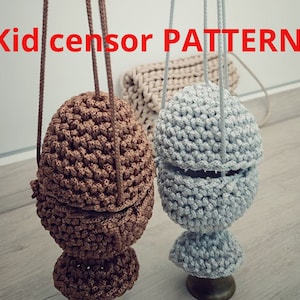 May include: Two handmade, crocheted egg-shaped items with long cords for hanging. One is brown, the other is gray. The text "Kid censor PATTERN" is in red at the top. The items are suspended in front of a neutral background.